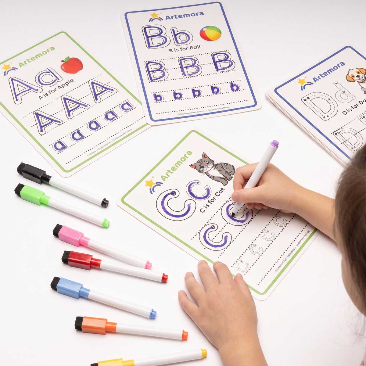Artemora alphabet tracing sheets for children shown on a white surface, with a child's hand writing on a green letter C card and several coloured markers arranged nearby.